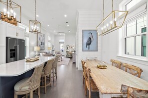 A spacious kitchen with a large island, wooden dining table, and chairs. There is a refrigerator, cabinets, and a painting on the wall.