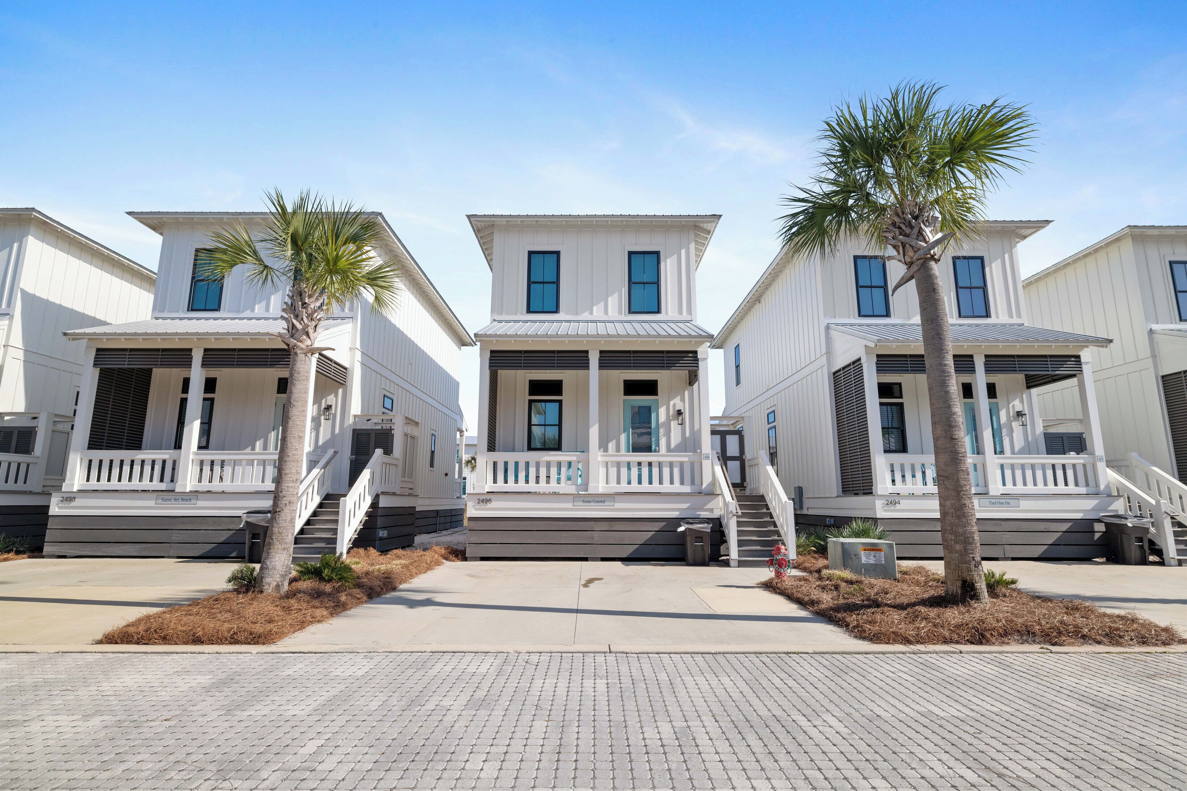A row of white two-story houses with a central entrance, each with a small porch and a palm tree in front.