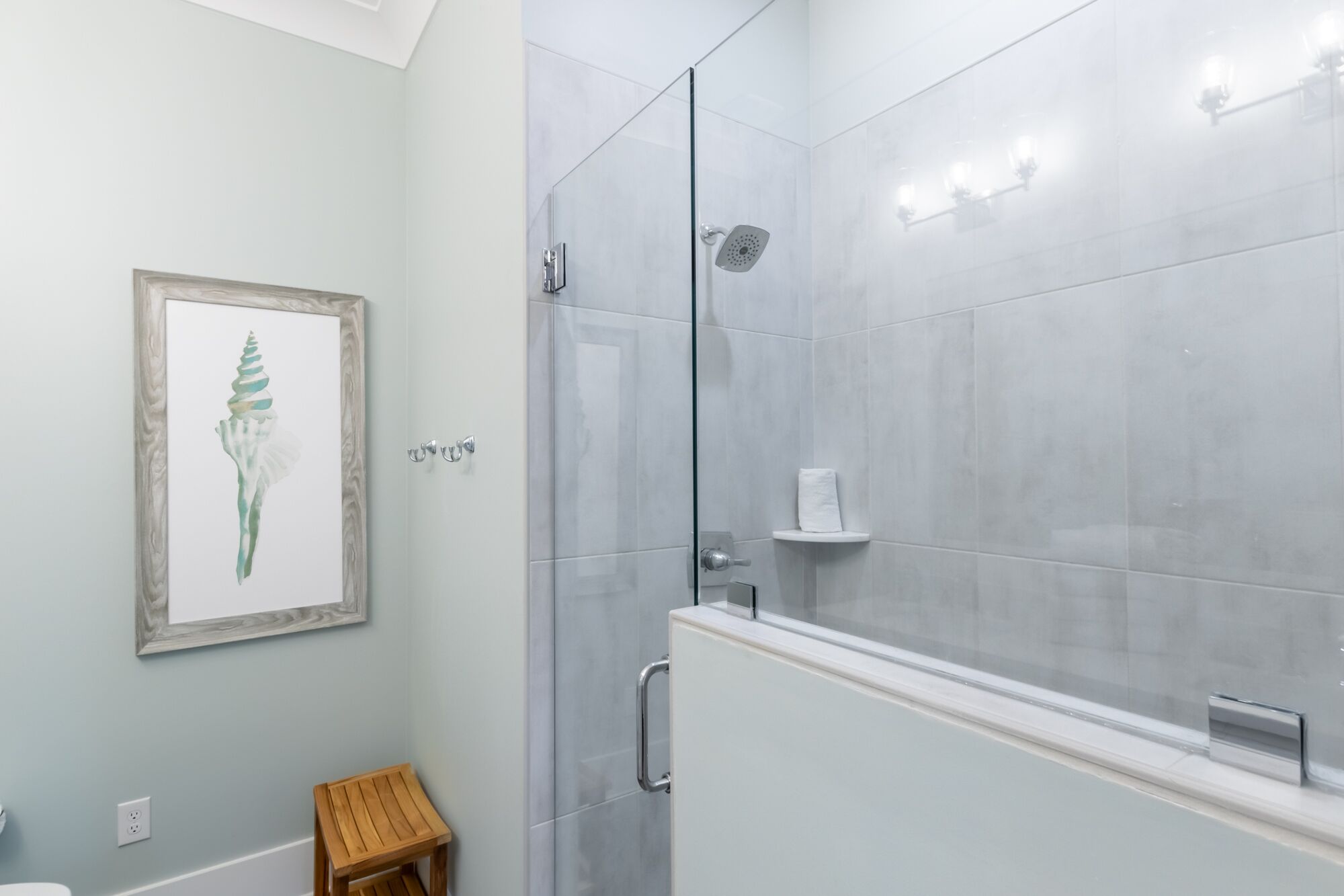 A modern bathroom with a glass shower enclosure, a wooden stool, and a framed artwork on the wall.