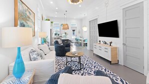 Una sala de estar moderna con un sofá, una mesa de centro y un televisor. Se ve una cocina al fondo con un comedor.