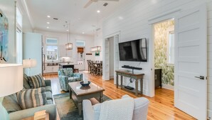 A spacious living room with a wooden floor, a dining area with a table and chairs, a kitchen with white cabinets, and a large flat-screen TV mounted on the wall.