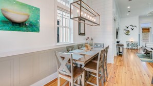 A dining area with a long table and chairs, a hanging light fixture, and a window with blinds.