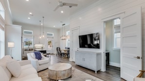 A spacious living room with a large TV, white furniture, and a dining area with a kitchen in the background.