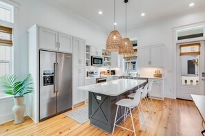 Uma cozinha moderna com armários brancos, eletrodomésticos de aço inoxidável, uma ilha grande com banquetas e um pendente de rattan.
