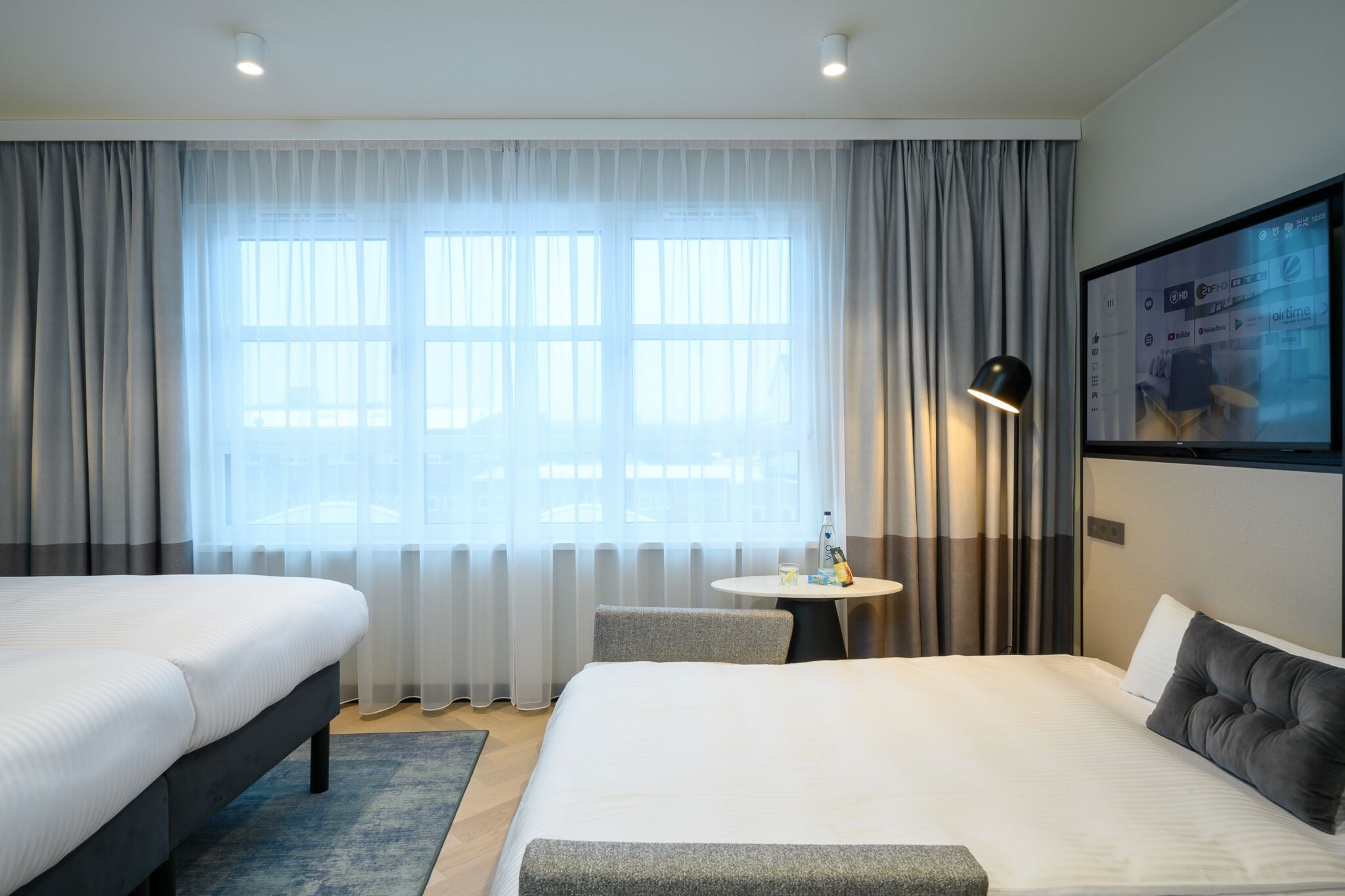 Ein Hotelzimmer mit zwei Betten, einem kleinen runden Tisch, einem an der Wand montierten Fernseher und einem Fenster mit durchsichtigen Vorhängen.