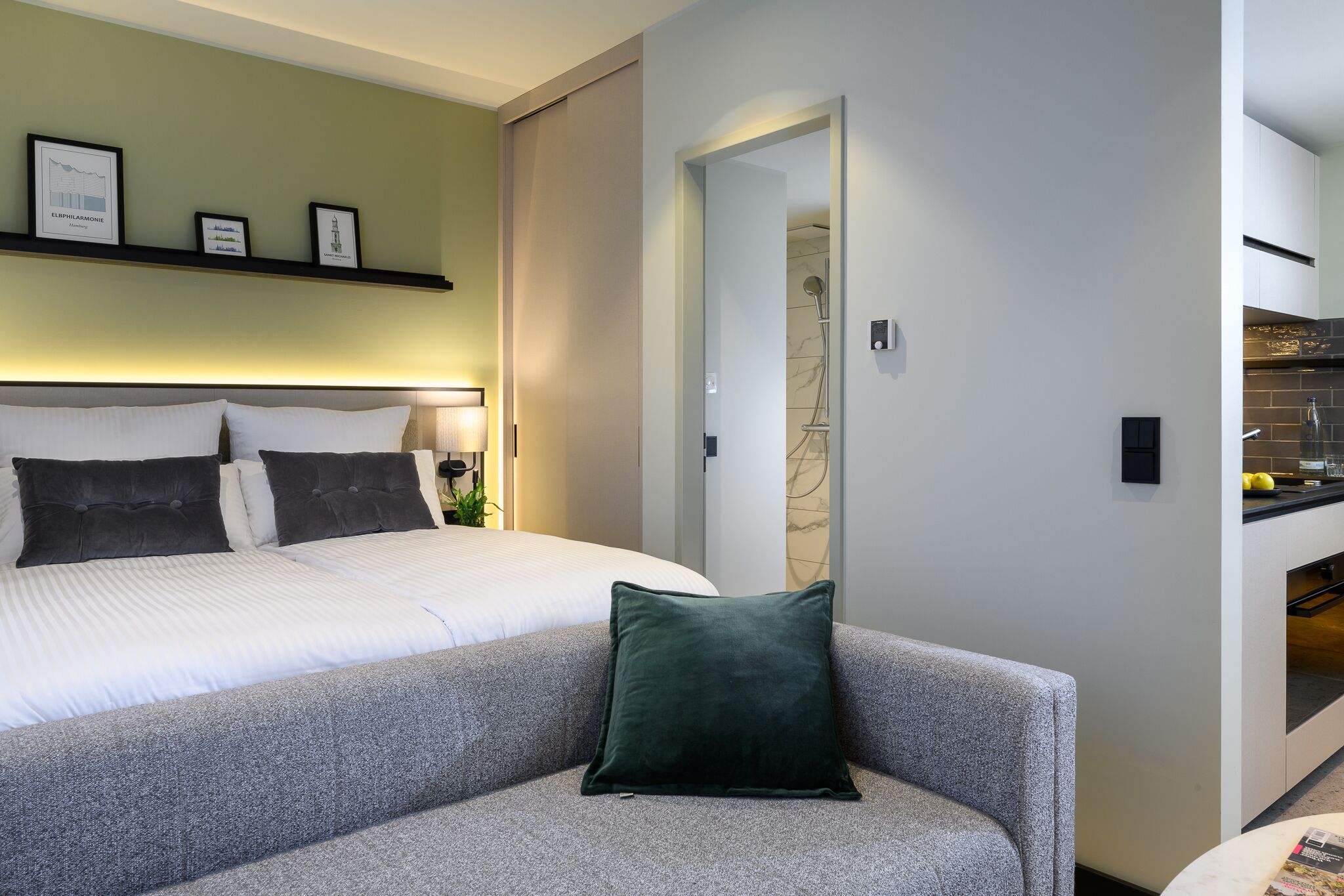 Een moderne hotelkamer met een net opgemaakt bed, een grijze bank met een groen kussen, en een keukenruimte zichtbaar door een geopende deur.