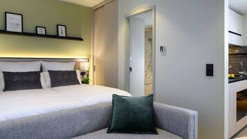 Een moderne hotelkamer met een net opgemaakt bed, een grijze bank met een groen kussen, en een keukenruimte zichtbaar door een geopende deur.