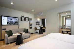 Una habitación de hotel moderna con una cama grande, un sofá, una pequeña mesa redonda y un televisor que muestra un menú de servicio a la habitación.