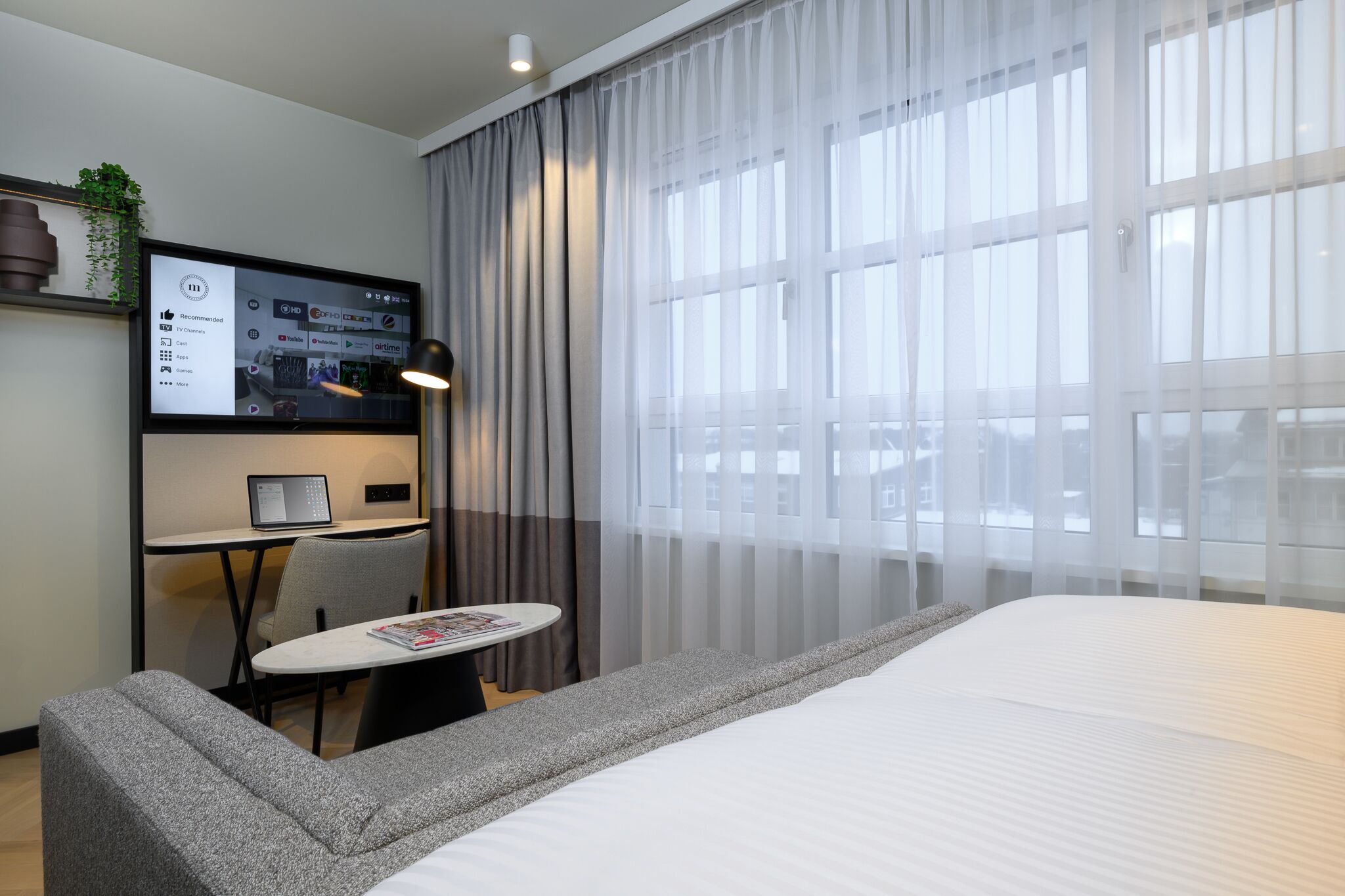 A modern hotel room with a large bed, a desk with a laptop, a small table with a magazine, and a TV displaying a hotel channel.