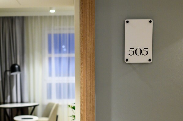 A room with a number sign '505' on the wall, a window with curtains, a lamp, and a small table.