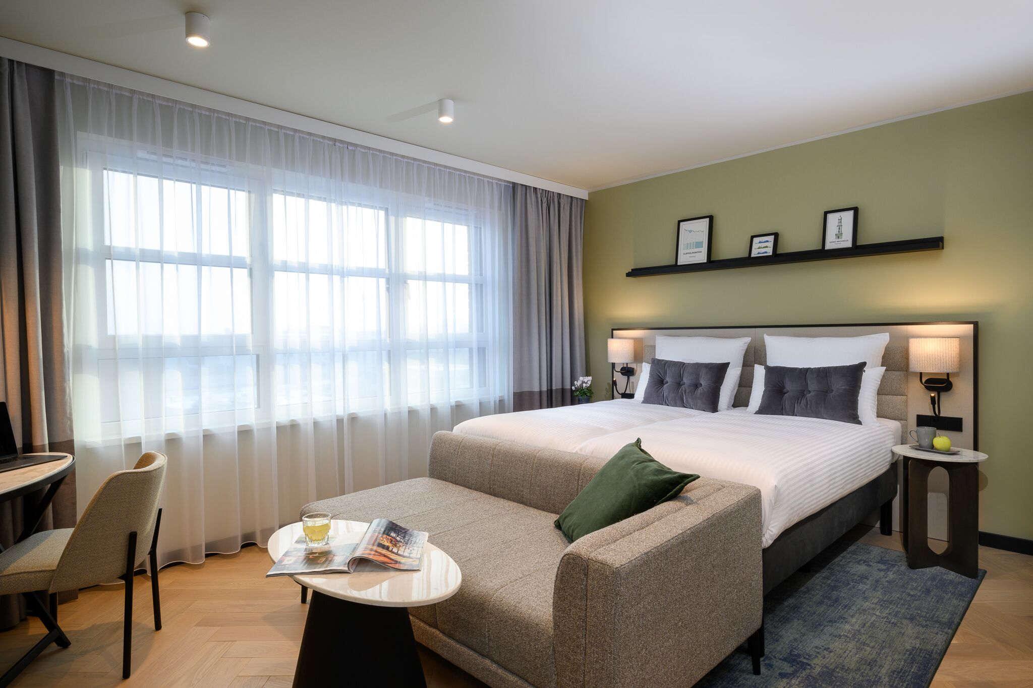A hotel room with a large bed, a sofa, a small table with a magazine, a desk, and a chair. There are two bedside lamps and a shelf with framed pictures.