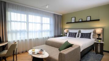 A hotel room with a large bed, a sofa, a small table with a magazine, a desk, and a chair. There are two bedside lamps and a shelf with framed pictures.