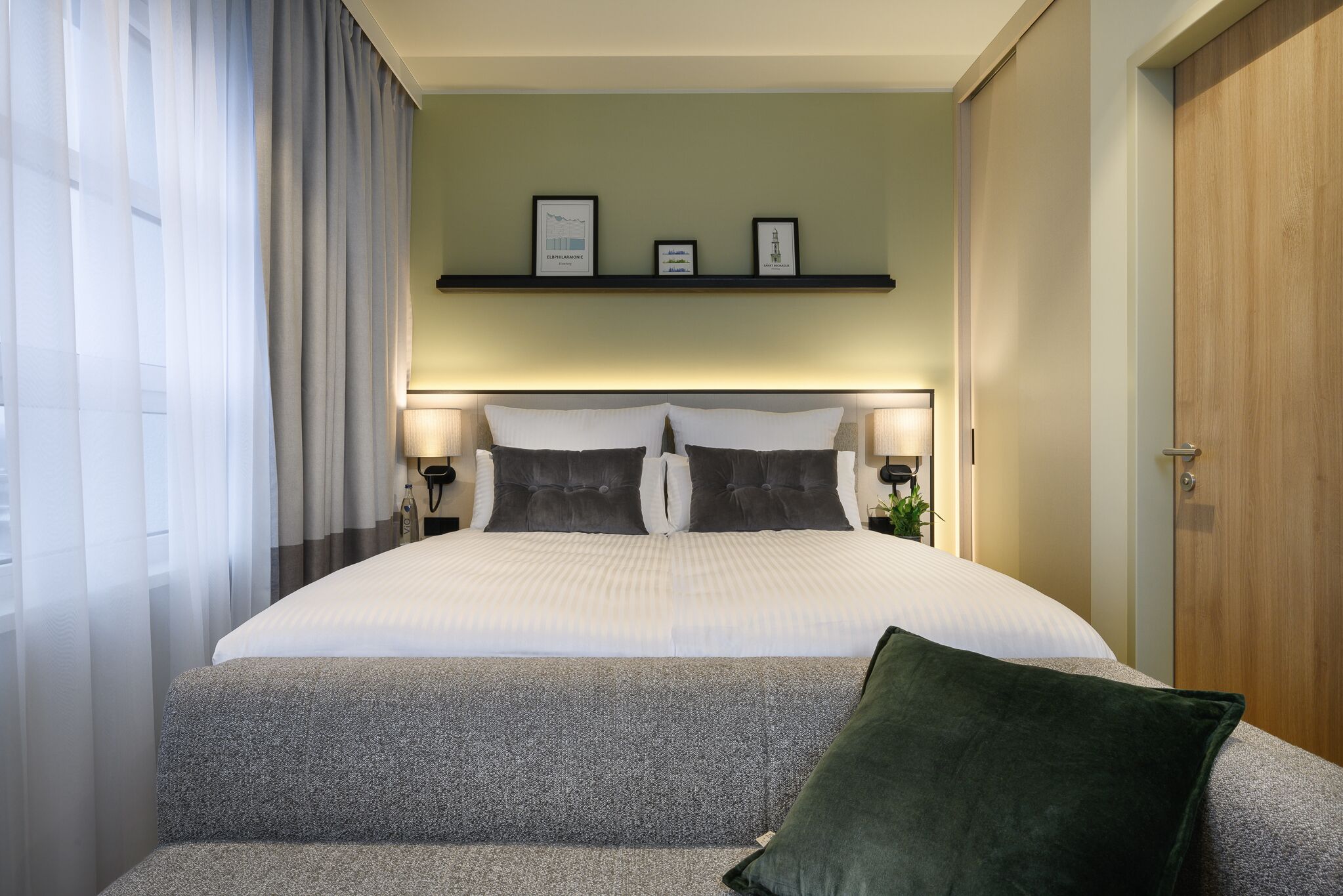 Une chambre d’hôtel bien aménagée, avec un grand lit, deux tables de chevet avec des lampes, une étagère avec des photos encadrées et un canapé vert recouvert de tissu.