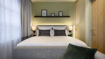 Une chambre d’hôtel bien aménagée, avec un grand lit, deux tables de chevet avec des lampes, une étagère avec des photos encadrées et un canapé vert recouvert de tissu.