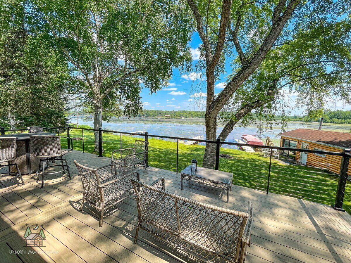 A deck with wicker furniture overlooking a lake, a wooden cabin, and a grassy area.