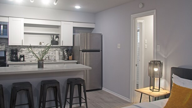 A modern kitchen with a stainless steel refrigerator, a bar area with stools, and a small table with a lamp.