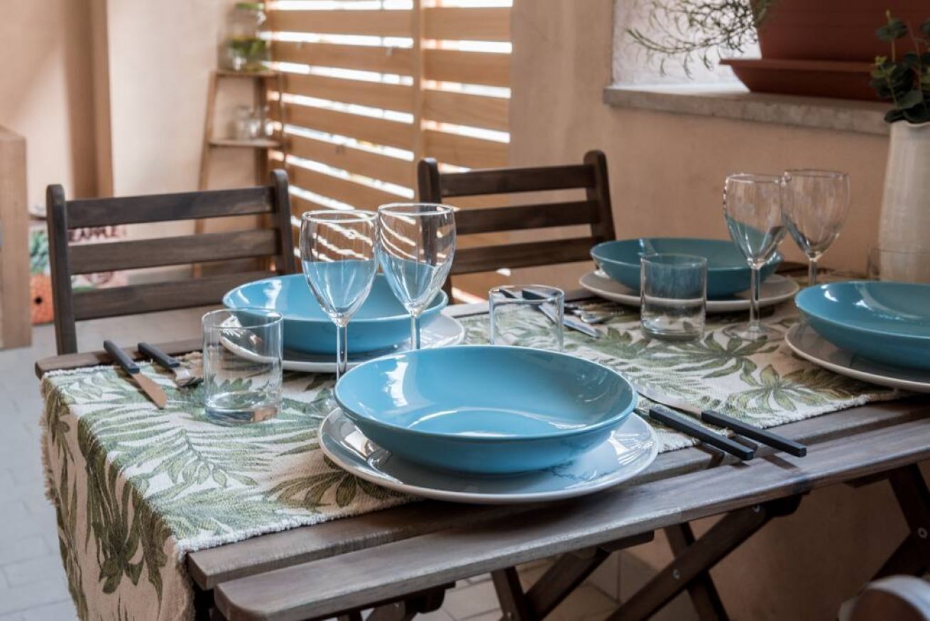 A dining table set with blue plates and clear glasses, a woven tablecloth, and wooden chairs.