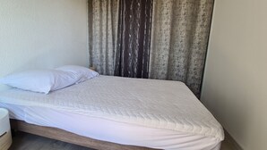 A neatly made bed with a white quilt, a bedside lamp, and a curtain. - F2 Confortable, à 2 Minutes de la Plage et du Port (CARNON-PLAGE)