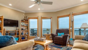 A living room with a large TV, wooden shelves, a brown armchair, and a view of the ocean through large windows. - New to Vrbo! "Dues Paid" Waterfront, Boat Dock, 3 Bedrm/2.5 Bath, Sleeps 6 (Dauphin Island)