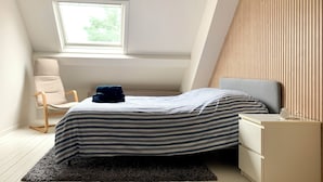 A bedroom with a sloped ceiling, a window with a view of greenery, a bed with striped bedding, a chair, and a bedside table with a lamp. - Cosy holiday home in Flanders Fields and at 10min from the sea. Pets welcome! (Alveringem)