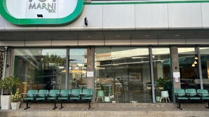 Uma loja Marni BKK com um logotipo verde e branco, grandes janelas de vidro e área externa com plantas em vasos.