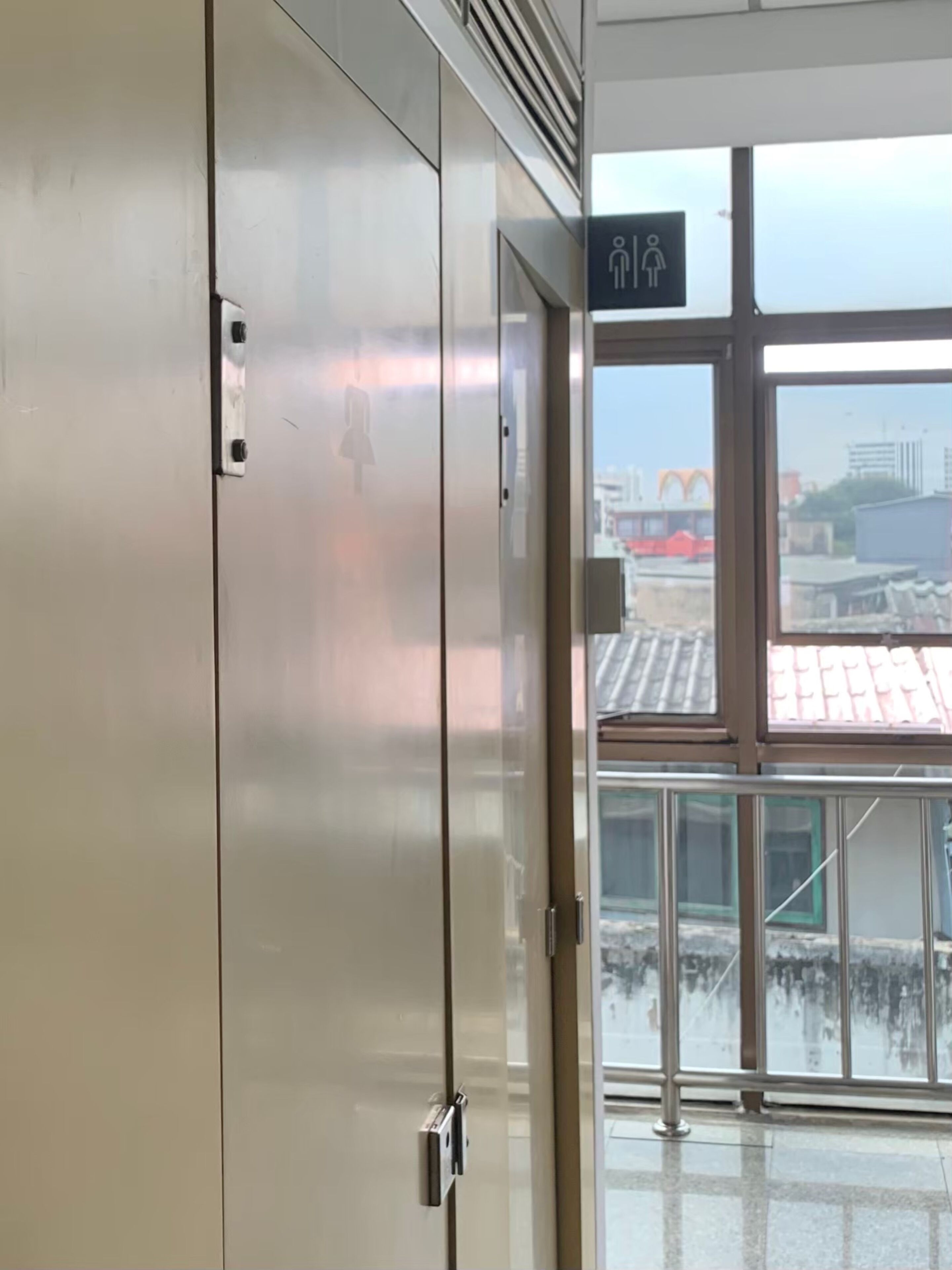 A modern elevator with a glass door, a visible emergency exit sign, and a view of the cityscape through the windows.