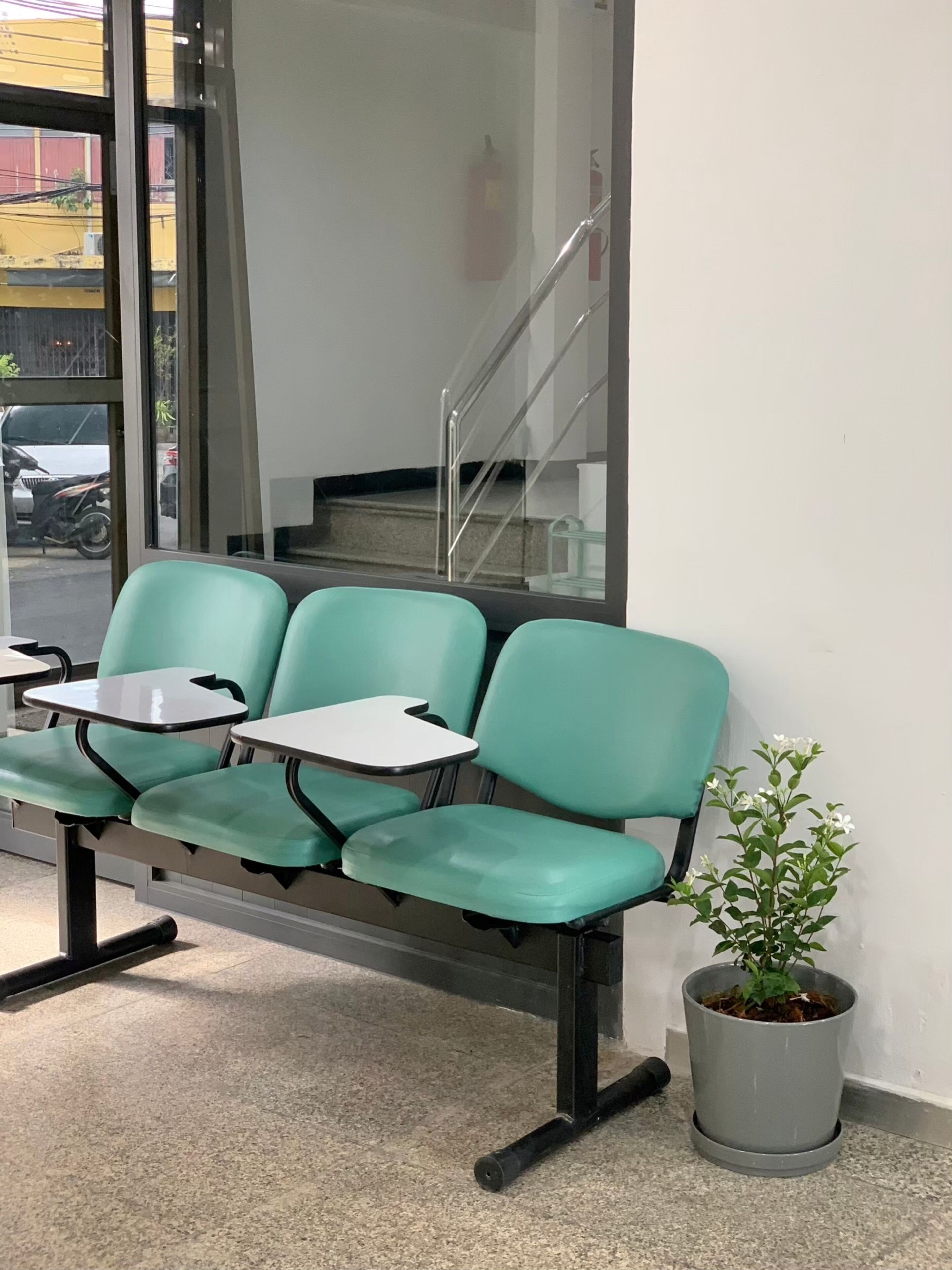 A waiting area with three green chairs and small tables, a potted plant, and a fire extinguisher on the wall.