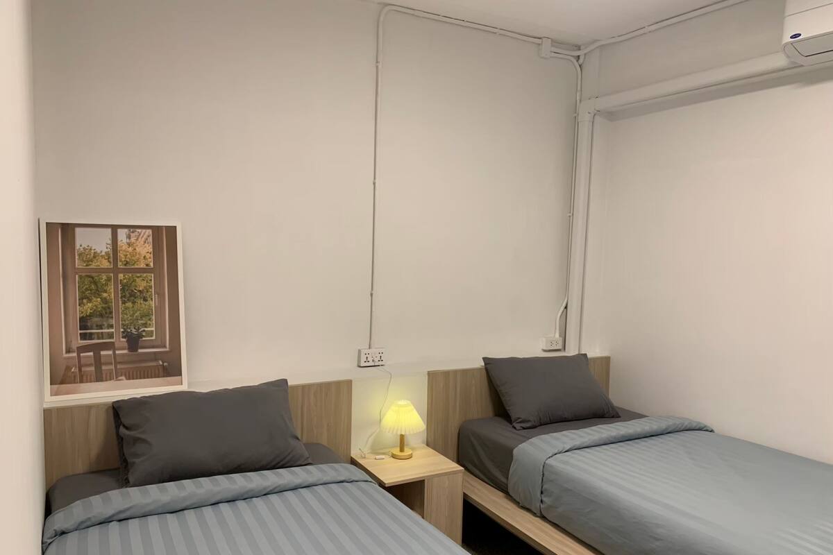 A bedroom with two single beds, a nightstand with a lamp, and a window with a view of greenery.