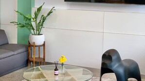 Uma sala de estar com uma mesa de centro redonda de vidro, uma poltrona em formato de elefante preto, uma planta em vaso e uma televisão de tela plana na parede.
