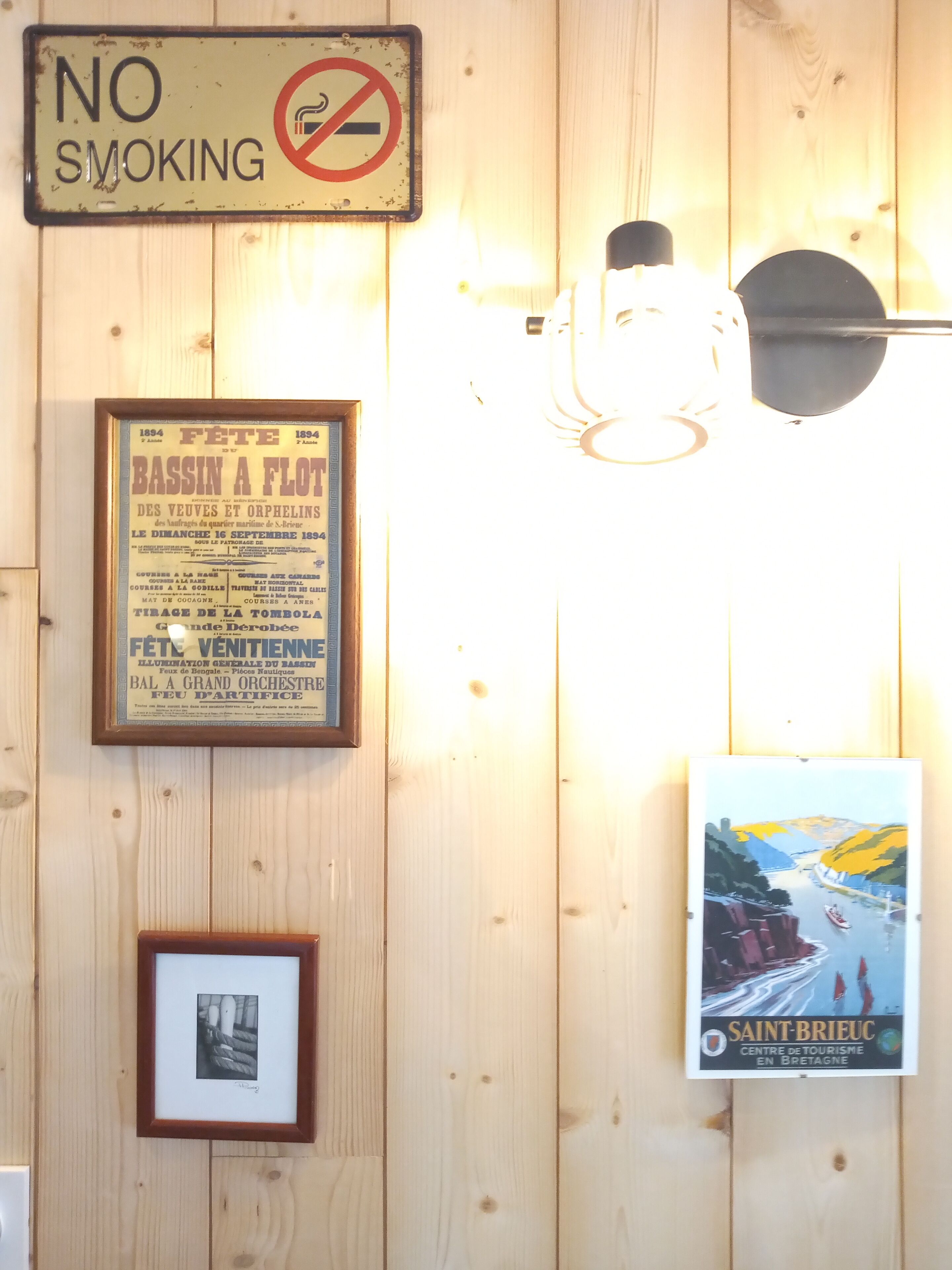 Un mur orné d’un panneau « Il est interdit de fumer », d’une affiche ancienne encadrée, d’une photographie encadrée et d’une affiche pour l’« Office de tourisme de Saint-Brieuc en Bretagne ».
