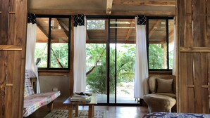 A wooden room with a bed, a sofa, and a table. There are large windows with curtains. - Playa Pelada Beachfront Bungalow at villas Ecositas Nosara (Provincia de Guanacaste)