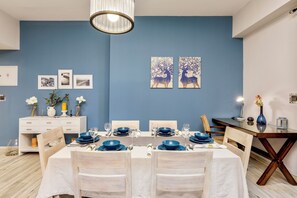 A dining area with a blue wall, white tablecloth, and blue tableware. There are framed pictures on the wall and a vase with flowers on the table.