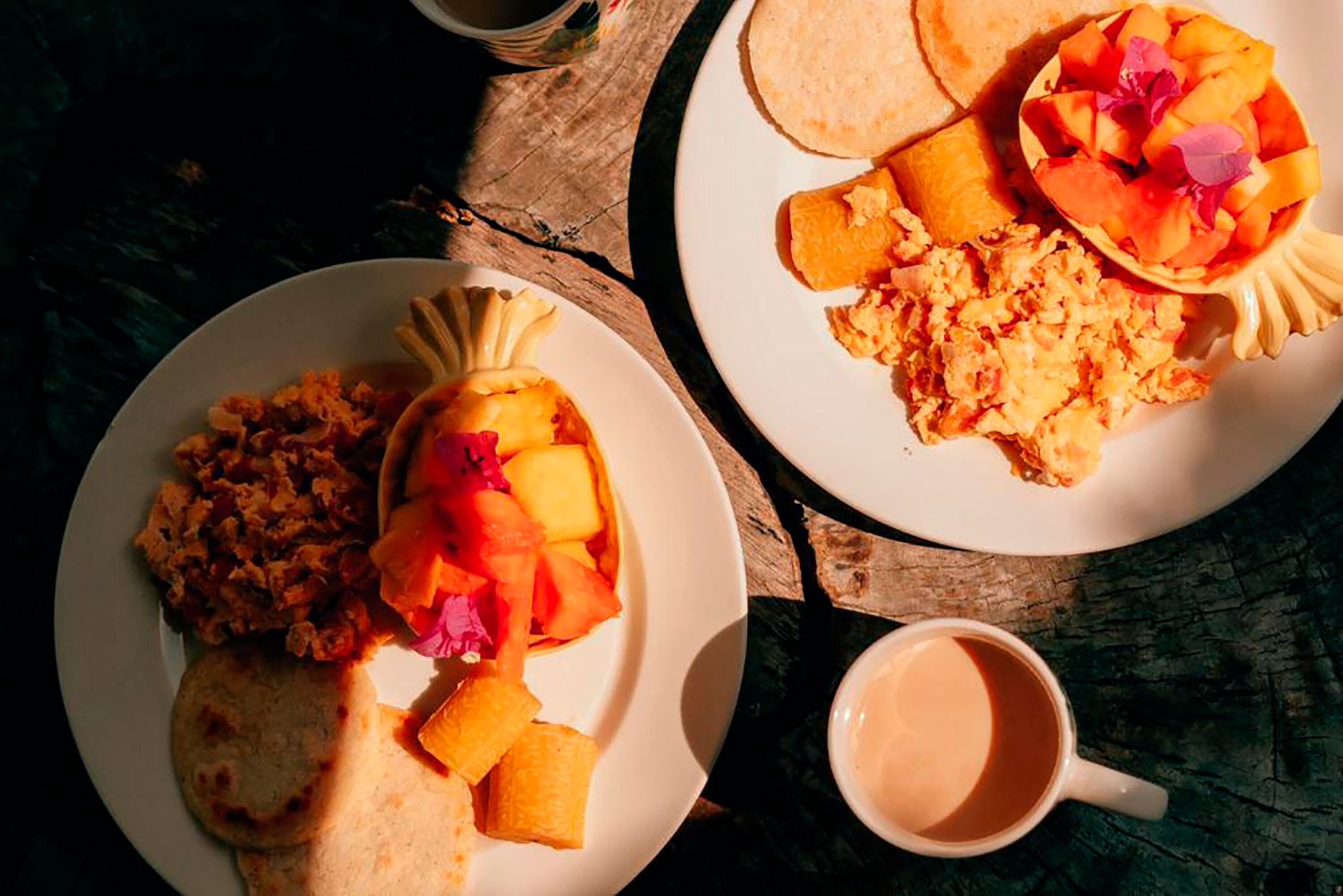 Dos platos de comida con fruta y huevos revueltos, una taza de café y una mesa de madera rústica.