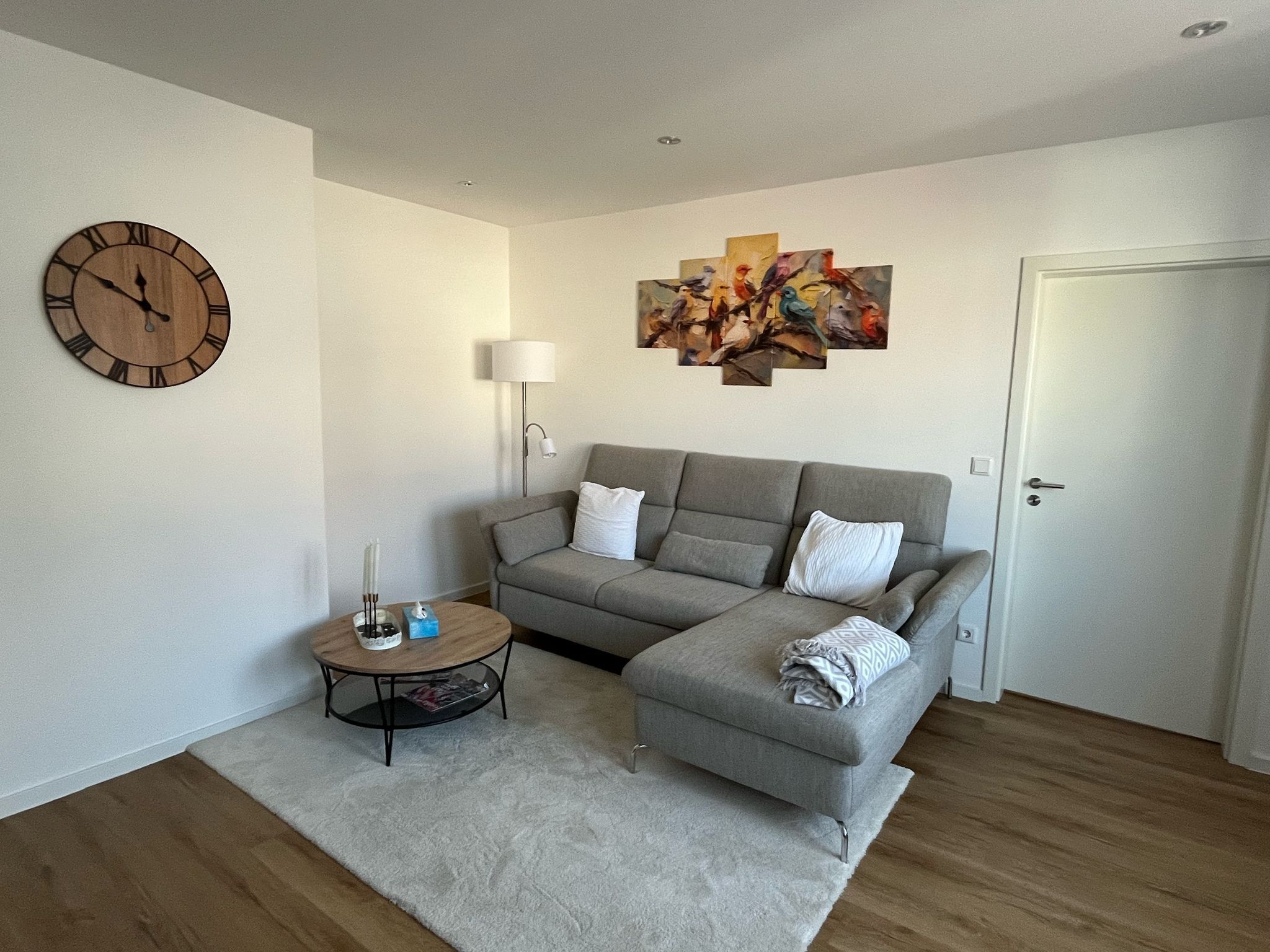 Una sala de estar moderna con un sofá seccional gris, una mesa redonda de centro, un reloj de pared, una lámpara de pie y un cuadro abstracto y colorido.