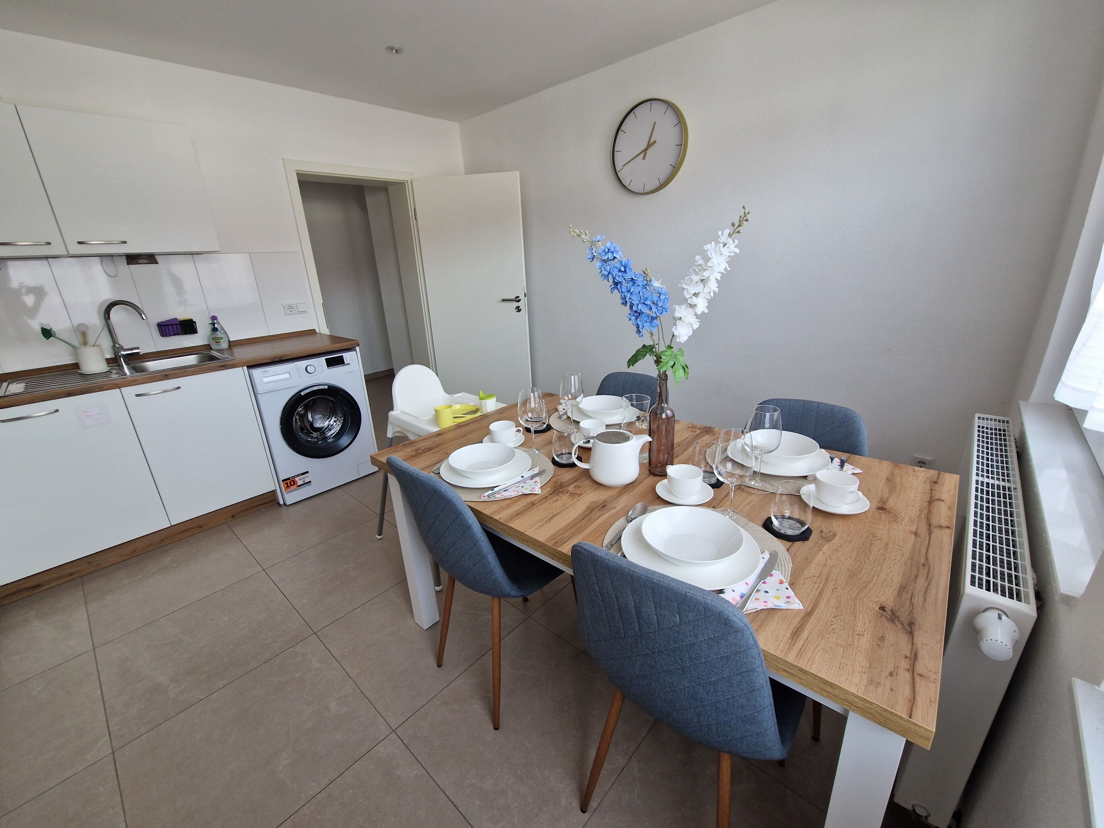 A modern kitchen with a washing machine, a dining table set with white plates, glasses, and a vase with flowers.