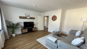 A modern living room with a grey sofa, a TV on a wooden stand, a potted plant, and a clock on the wall. - Bright, newly renovated TOP flat in a central location (Osnabrück)