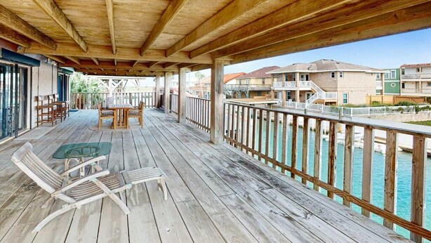 A wooden deck with a glass table and chairs, overlooking a marina with boats and buildings. - Channel-Front Port Isabel Family Home. Gulf/Bay Fishing and Close to SPI Beach! (Port Isabel)