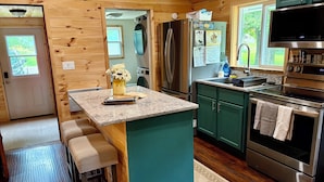 A compact kitchen with wooden walls, a central island, bar stools, a refrigerator, a stove, and a microwave. - Swimming, Snowmobiling, Serenity! Lakefront Cottage on Big Manistique Lake (McMillan)