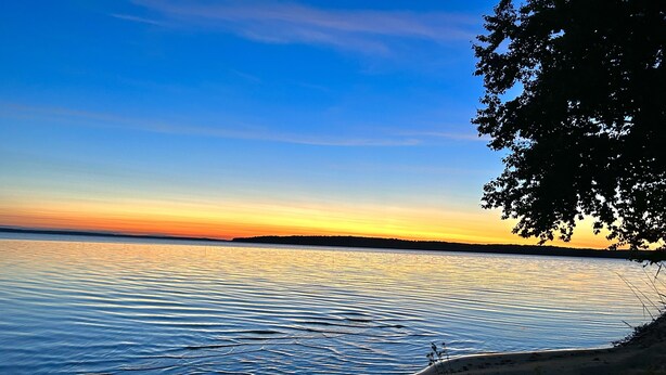 A beach with calm water, a clear sky, and a distant shoreline. - Swimming, Snowmobiling, Serenity! Lakefront Cottage on Big Manistique Lake (McMillan)