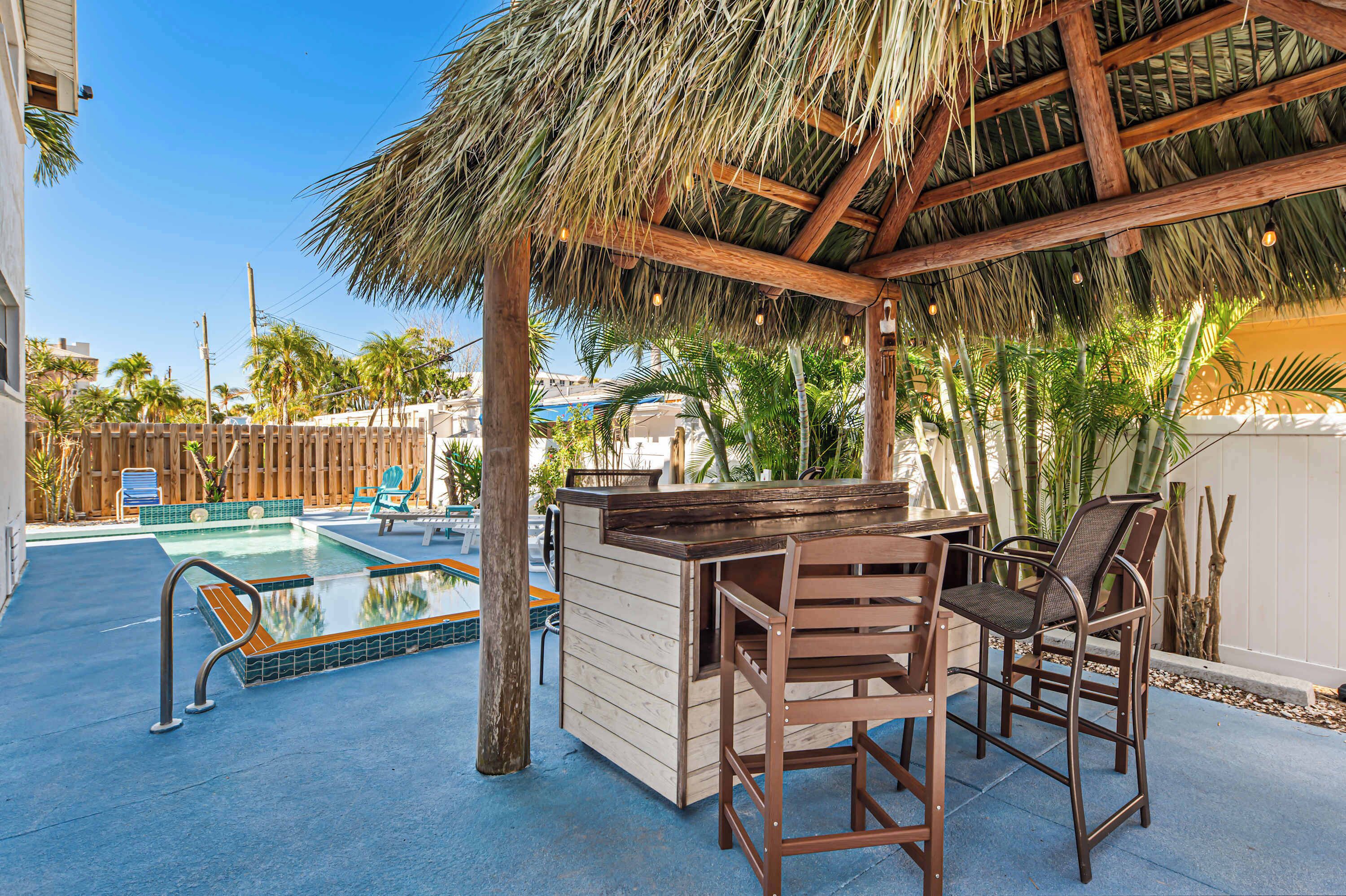 A pool area with a wooden bar and chairs, surrounded by palm trees and a thatched roof structure.