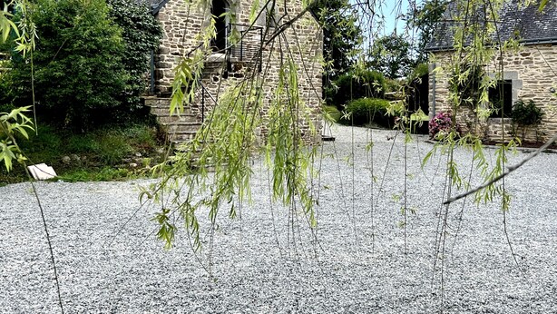 Un bâtiment en pierre avec une allée de gravier, entouré d’arbres et de verdure.