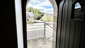 Une vue à travers une arche en pierre, donnant sur un balcon orné d’une balustrade en métal, qui surplombe un paysage rural avec des bâtiments en pierre et des arbres.