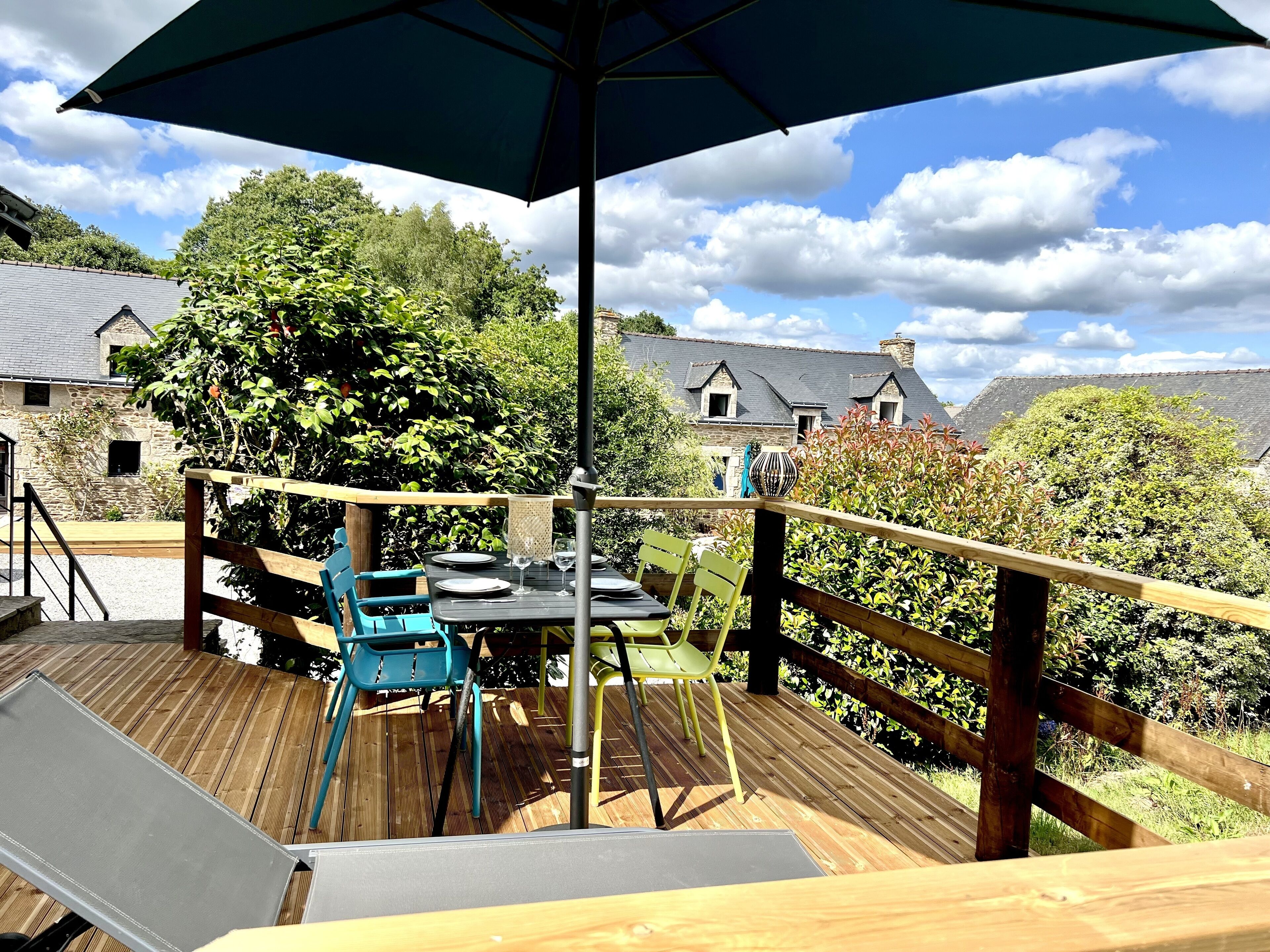 A wooden deck with a table and chairs, an umbrella, and a view of a stone building with a thatched roof.