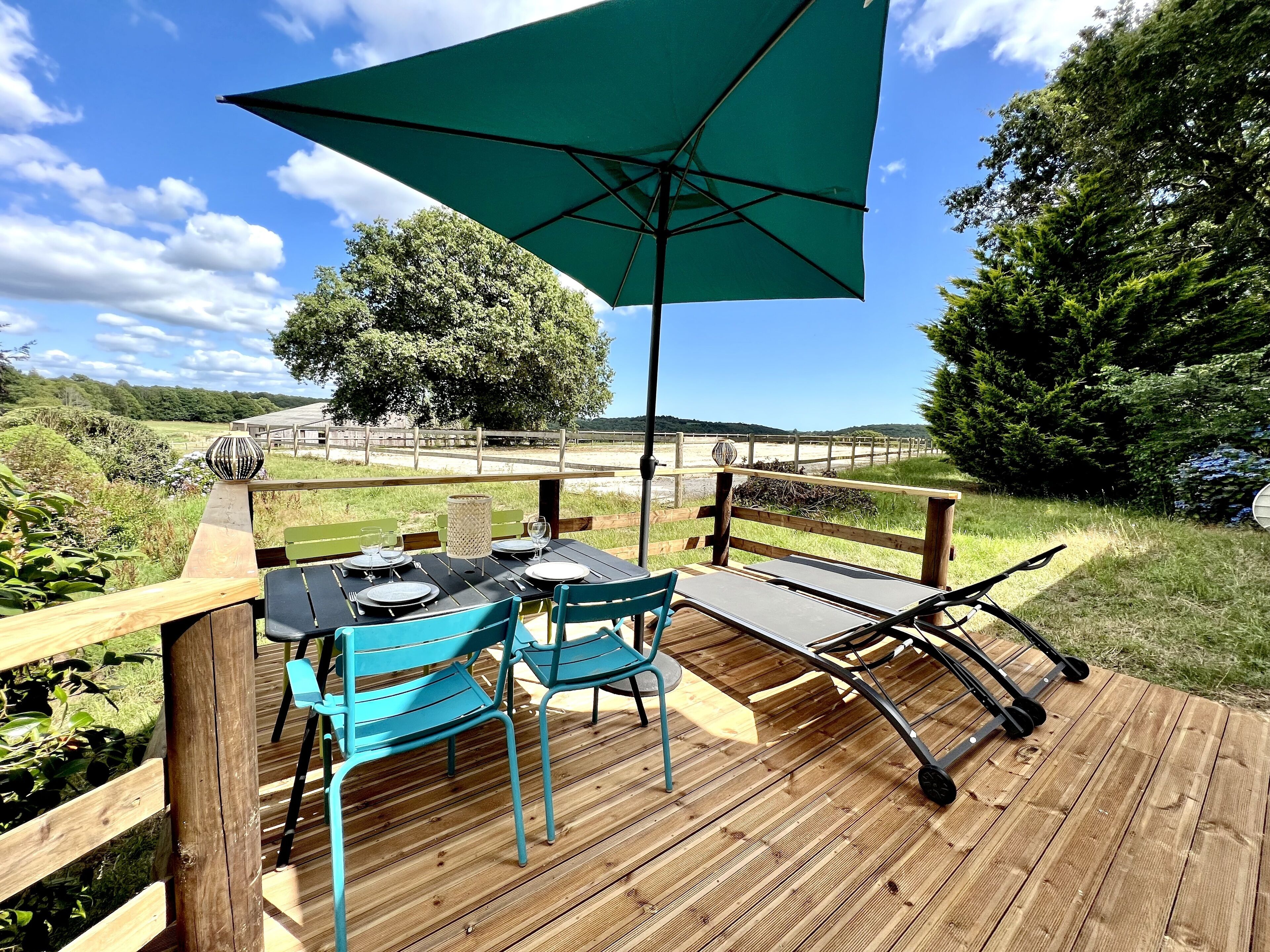 Eine Holzterrasse mit Tisch und Stühlen, einem großen Sonnenschirm und Blick auf Strand und Bäume.