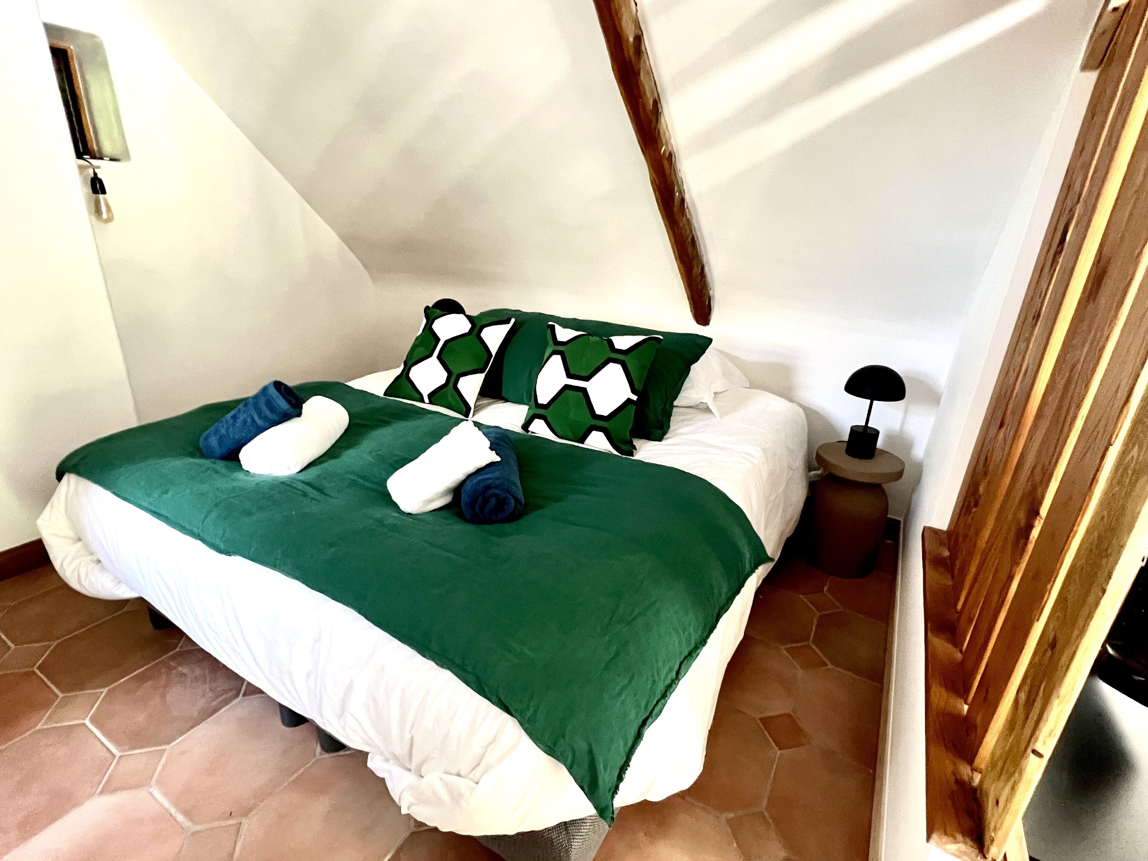 Una cama bien hecha con ropa de cama verde y blanca, un cabecero de madera y una mesita de noche con una lámpara.
