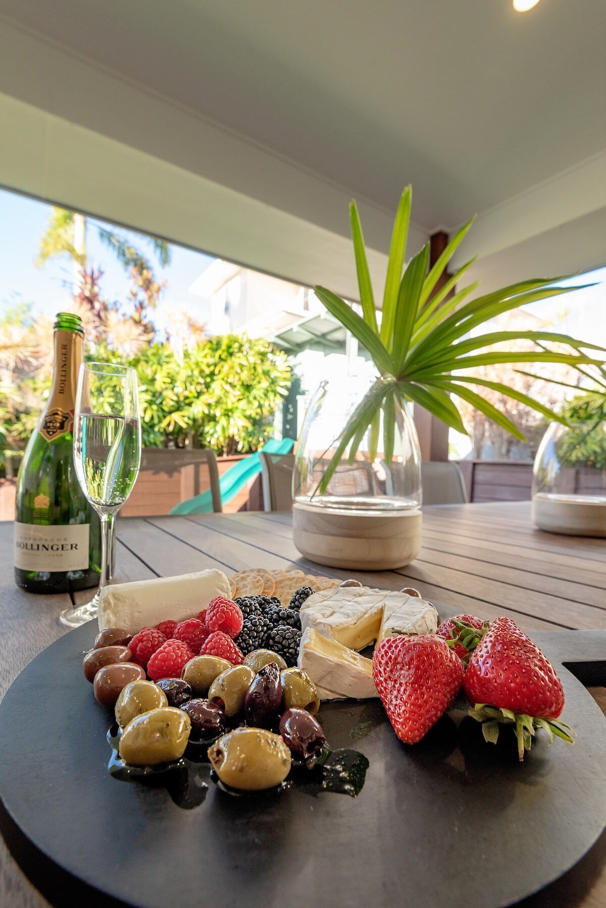 A table with a bottle of Bollinger champagne, a glass, a vase with a plant, and a charcuterie board with cheese, fruits, and olives.