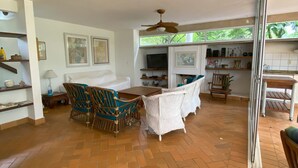 Un vaste espace de vie doté d’un parquet en point de Hongrie, d’une table à manger avec des chaises en osier, d’un ventilateur de plafond et d’un coin cuisine avec une fenêtre donnant sur la verdure.