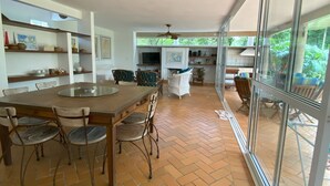 Une cuisine spacieuse avec une grande table à manger, des chaises et un téléviseur. On y trouve des étagères remplies de divers objets et une fenêtre donnant sur l’extérieur.