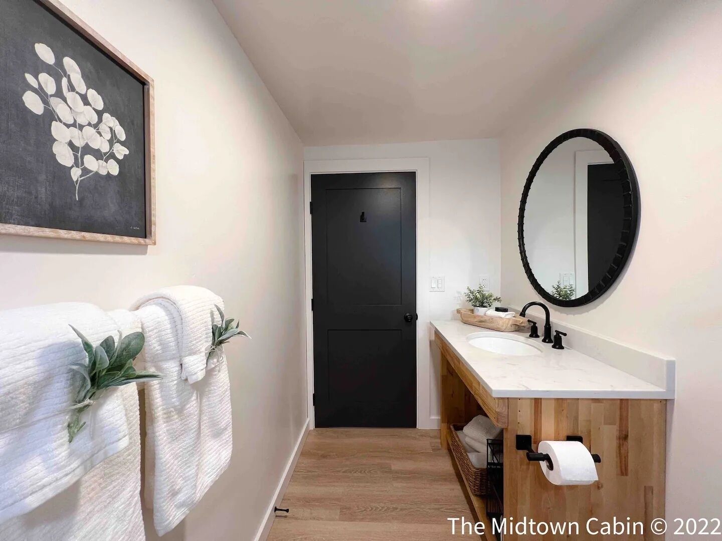 A modern bathroom with a wooden vanity, a black door, and a framed artwork on the wall.