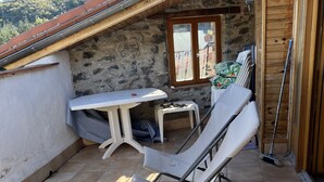 Eine sonnendurchflutete Terrasse mit einem Metallstuhl, einem Tisch und einem Fenster mit Blick auf eine bewaldete Landschaft.